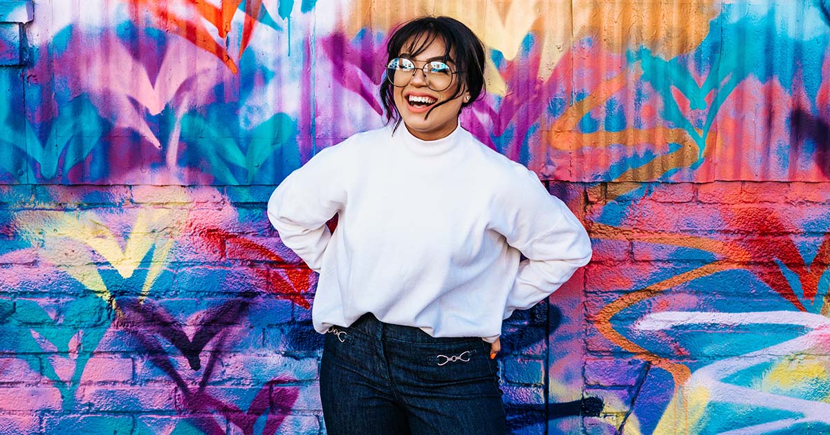 Woman in front of a graffiti wall in a white sweater and glasses smiling